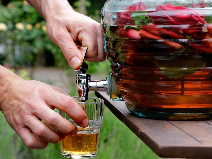 En person skænker sig et glas saft fra en stor glas-dispenser fuld af rabarbersaft og hele rabarberstykker i. 